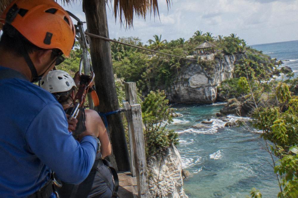 A guide in a helmet clipping a carabiner onto a rider's harness at a jungle zipline platform