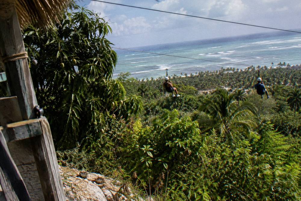 Two riders in helmets and harnesses gliding along parallel zipline cables above the forest