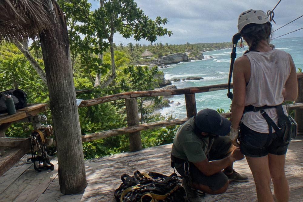 A guide checking a harness buckle on a wooden zipline platform before a rider launches