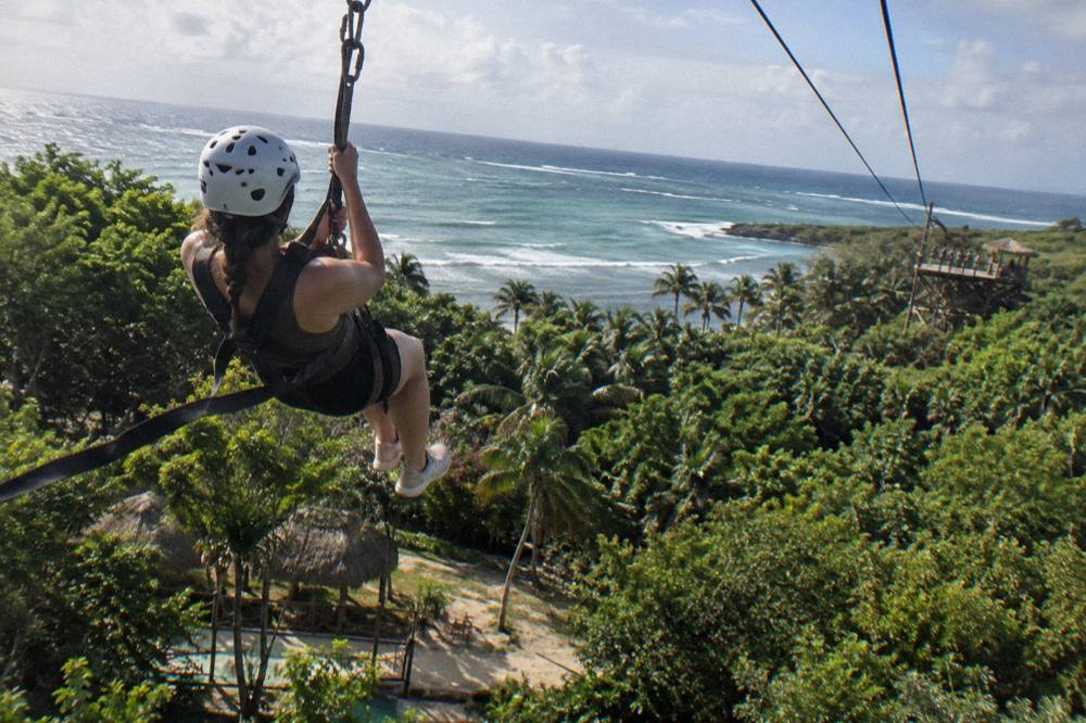 Zipline rider in a harness gliding above tropical treetops toward a distant wooden platform