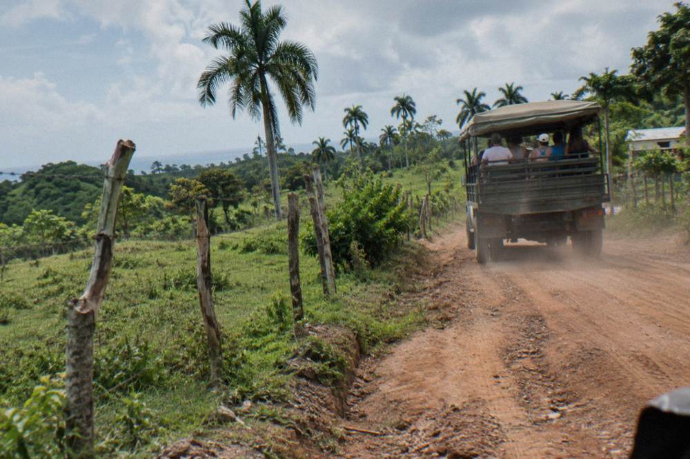 Open-sided safari truck carrying passengers along a dirt road through green mountain farmland