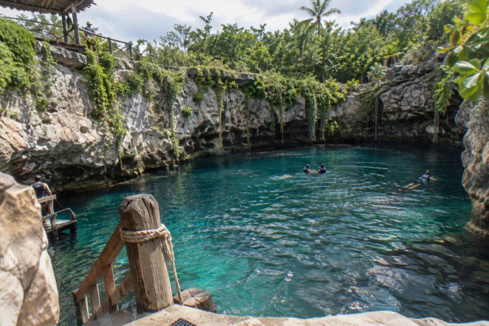 Turquoise cenote pool at the base of a limestone cliff surrounded by tropical vegetation