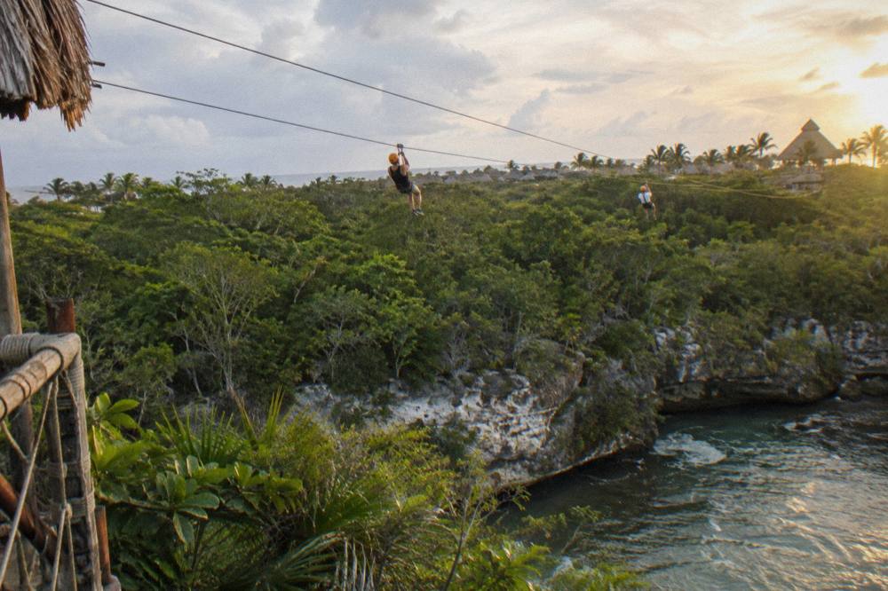 Two riders on parallel zipline cables crossing above a forested ravine