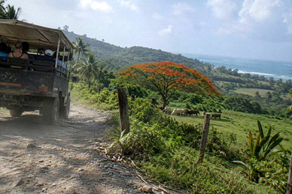 Safari truck driving on a rural road through green hills toward a mountain zipline base