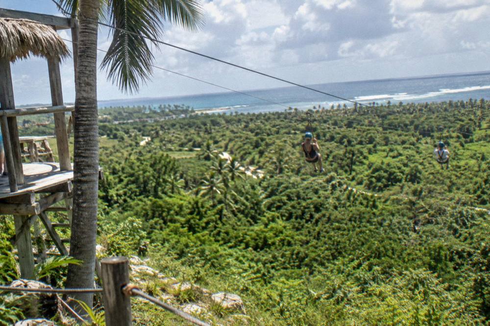 Visitors in helmets riding a zipline side by side over a jungle valley