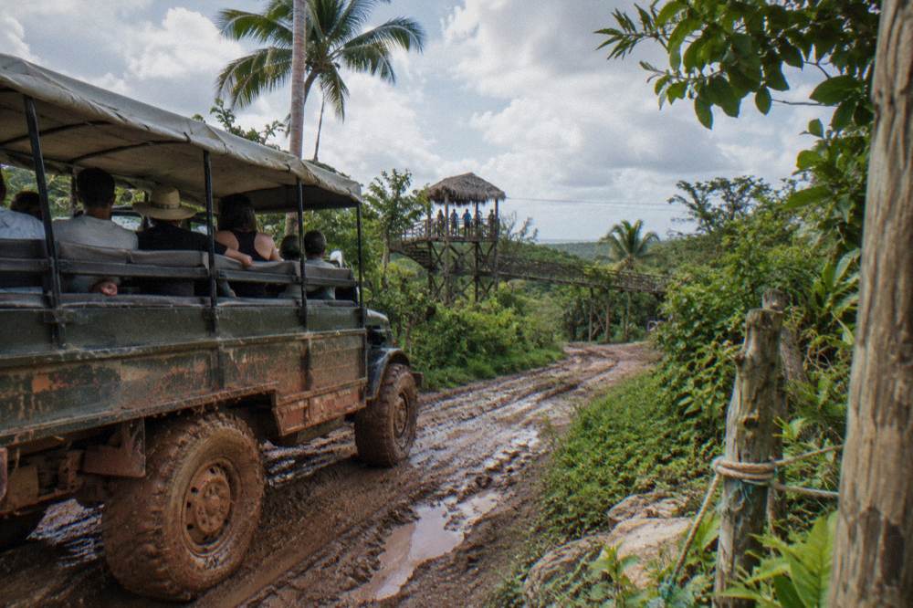 Open safari truck driving a jungle road toward mountain zipline platforms under broken clouds