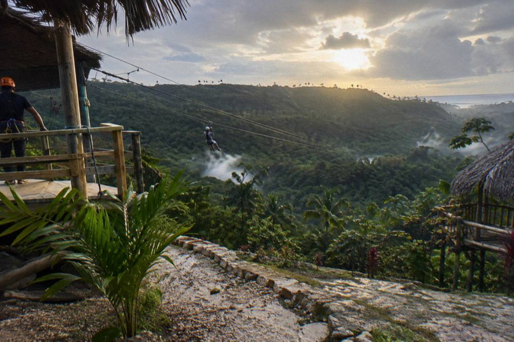 Sunlight breaking through clearing clouds over a tropical mountain ridge with zipline cables crossing a valley