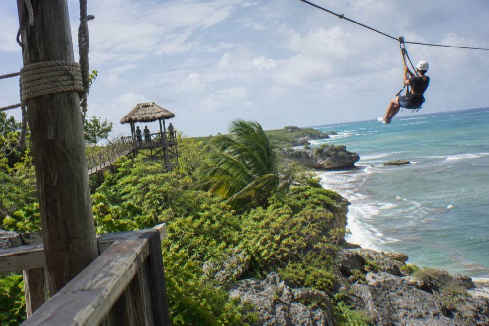 Person in helmet and harness clipped onto a steel zipline cable above forest canopy