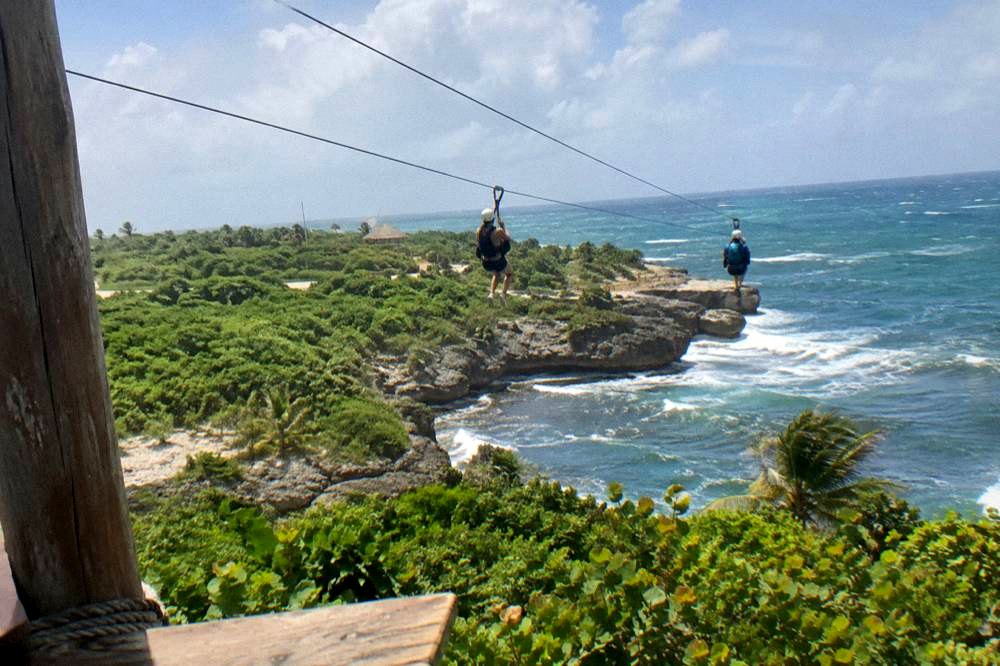 Two riders on parallel zipline cables above dense tropical forest