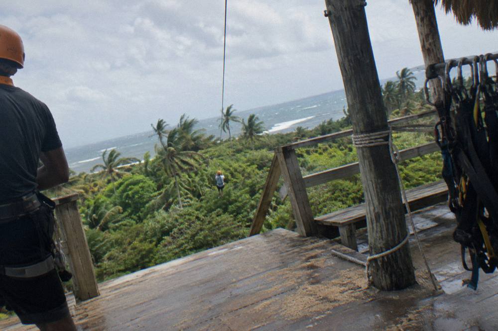 Child in helmet and harness standing on a wooden zipline platform beside a guide