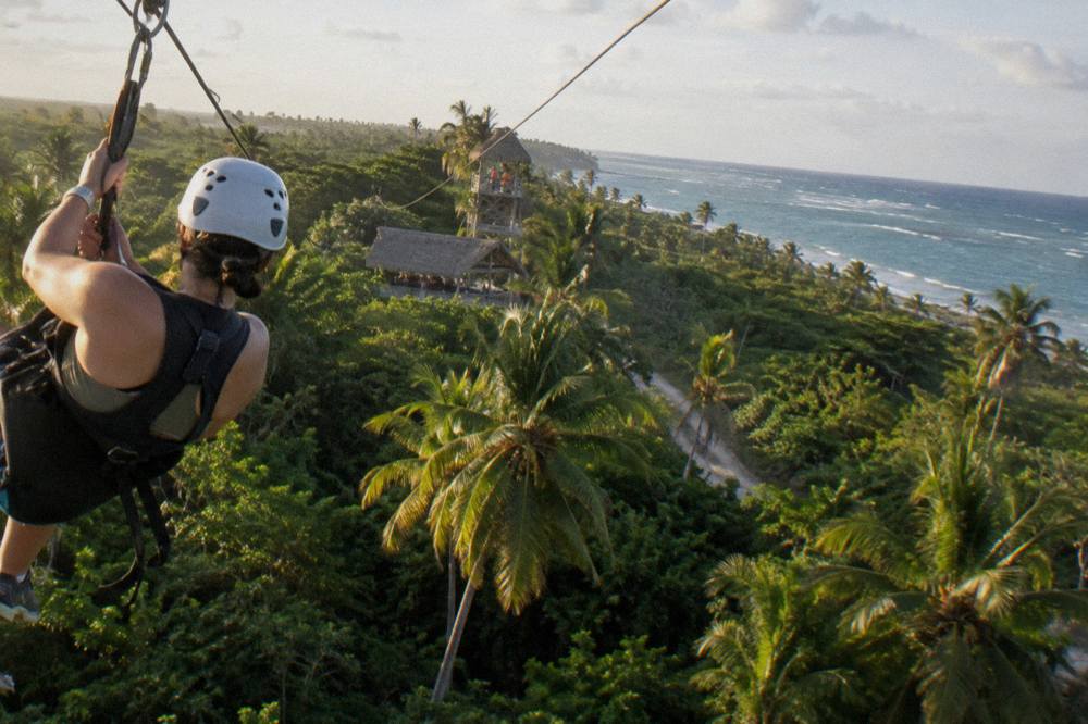 A zipline rider in a helmet and harness gliding above dense green jungle canopy.