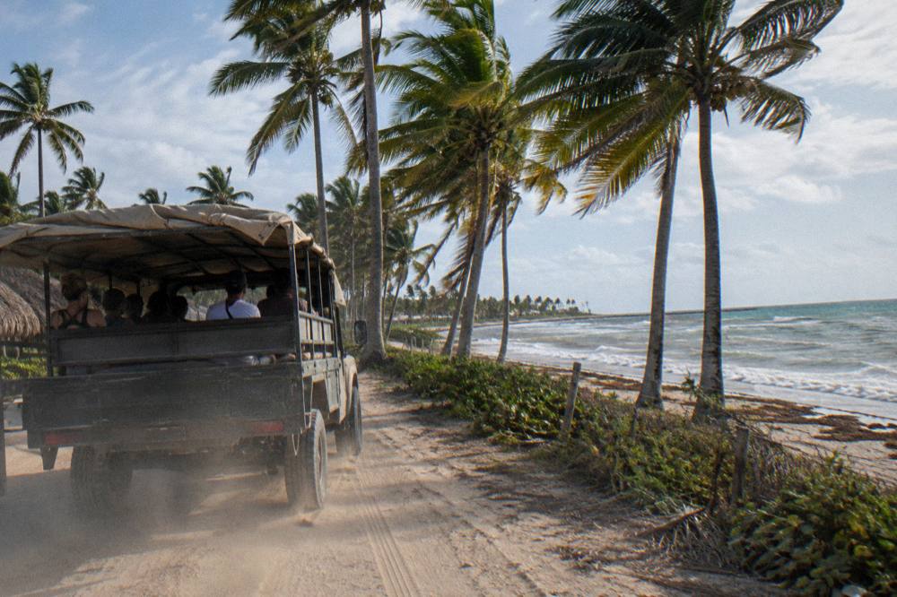 An open safari-style truck carrying zipline guests along a rural road past palm trees.