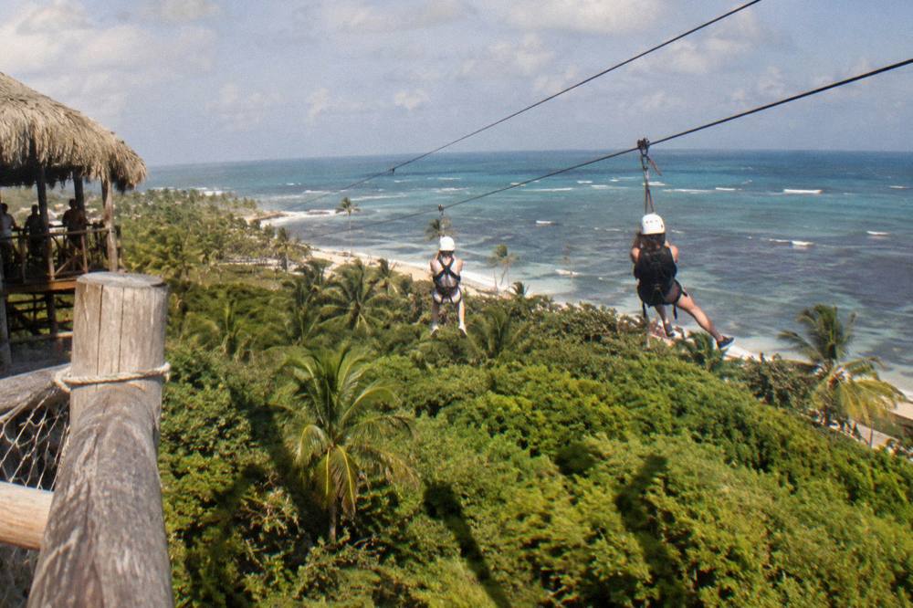 Two zipline riders on parallel cables gliding side by side above the treeline