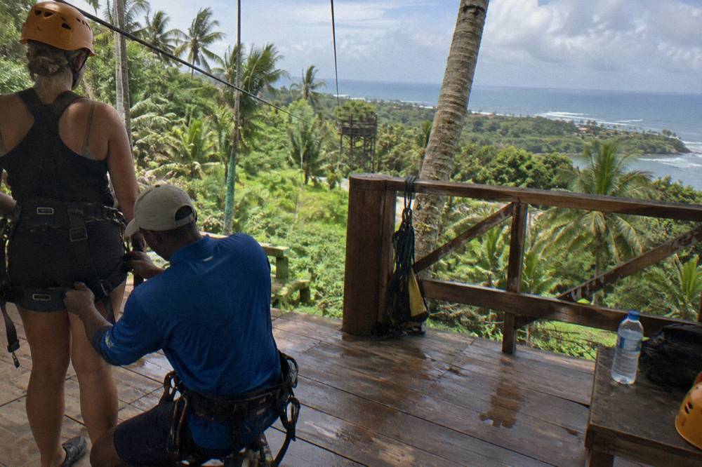 A guide checking harness straps on a wooden zipline platform surrounded by palm trees