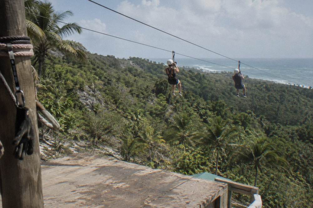 Two riders on parallel zipline cables crossing above a forested valley