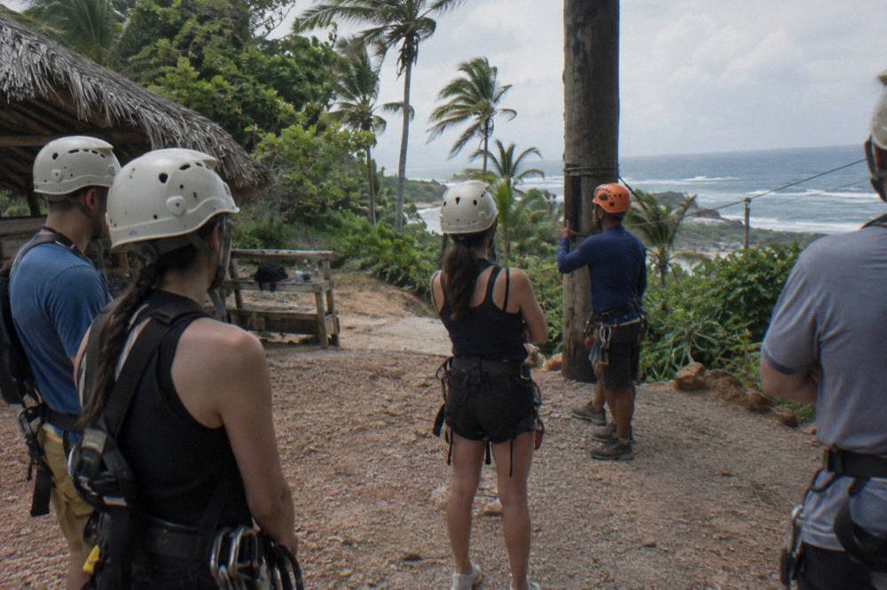A group of helmeted riders listening to a guide's safety briefing at a jungle zipline base
