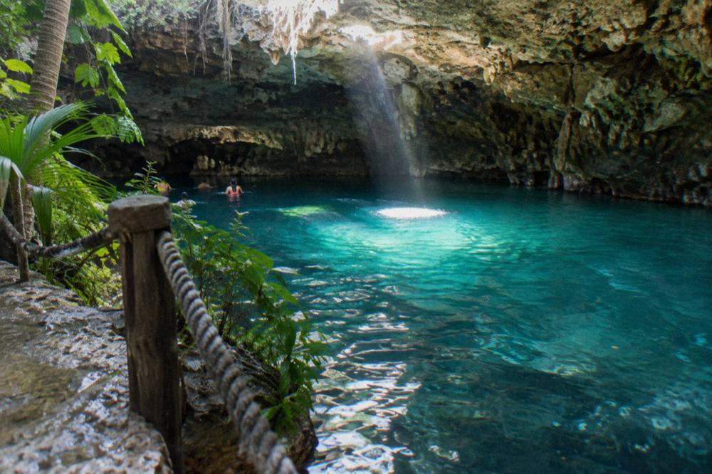 A cave cenote with still turquoise water lit by a shaft of daylight through an opening in the rock ceiling