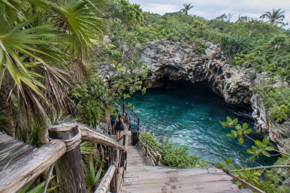 A wooden walkway leading through dense jungle toward a rock opening with water visible inside
