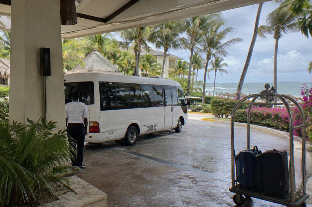 Covered resort arrival area with a shuttle minibus, luggage trolley, and tall palms